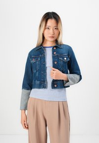 Denim jacket with a cropped design, blue color, orange buttons, chest pockets, grey sleeves. Worn over a striped shirt and beige trousers.