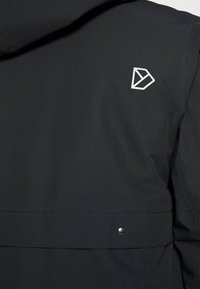 Črno zunanjo oblačilo z gladko, mat površino. Na hrbtni strani je prikazan bel geometrijski logo in zapenjanje s pritiskom na robu.