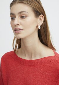 Boucles d'oreilles en forme de goutte d'eau en argent avec une surface réfléchissante ; portées avec un pull en tricot rouge présentant un décolleté large et une texture subtile.