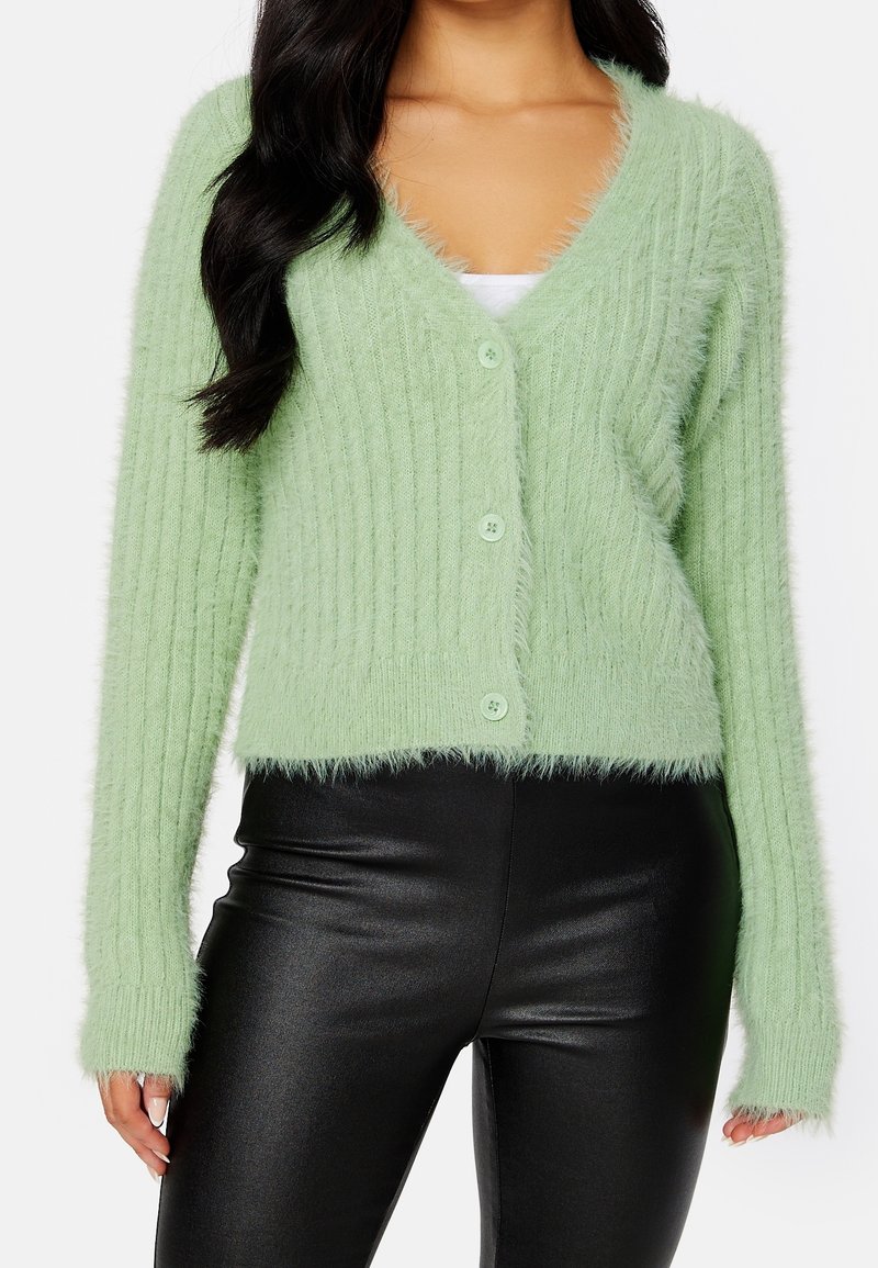 Femme portant un cardigan duveteux vert clair avec des boutons sur un haut blanc et un pantalon en cuir noir, montrée de la moitié de la poitrine jusqu'aux hanches.