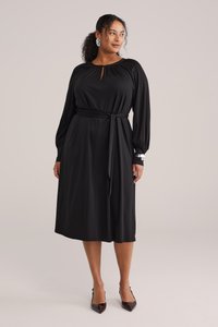 Schwarzes Kleid mit langen Ärmeln, Ausschnitt mit Schlitz, Taillengürtel. Hergestellt aus weichem Stoff, mit einer fließenden Silhouette und kontrastierenden Manschetten.