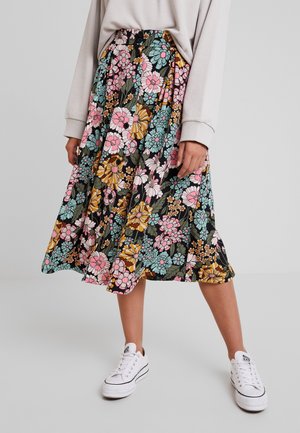 Personne portant des baskets blanches et une jupe midi fleurie colorée avec des fleurs roses, bleues et jaunes, associée à un haut à manches longues beige clair.