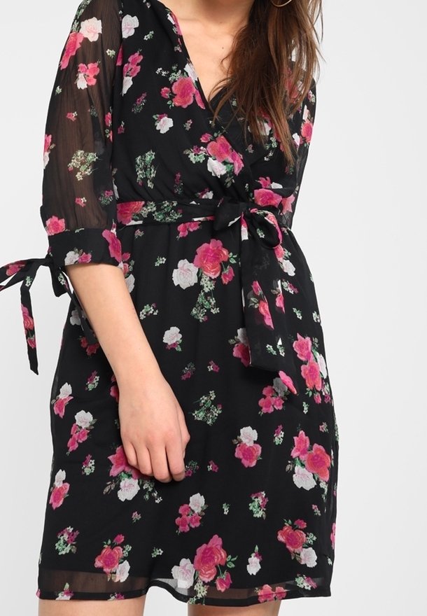 Kvinna klädd i en svart omslagsklänning med rosa och vita blommor, genomskinliga trekvartsärmar och knytning i midjan samt ärmar.