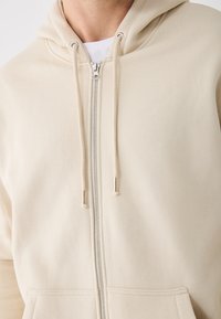 Bež zip-sv jumper s mešavinom mekanog pamuka, prednjim džepom, podesivom kapuljačom i srebrnim detaljima.