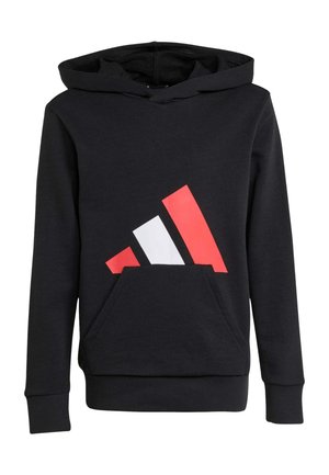Niño sonriente con sudadera con capucha y pantalones negros con un diseño de triángulo rojo y blanco sostiene una bolsa negra, de pie sobre un fondo liso y claro.
