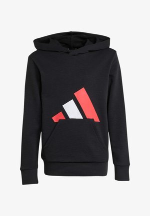Niño sonriente con sudadera con capucha y pantalones negros con un diseño de triángulo rojo y blanco sostiene una bolsa negra, de pie sobre un fondo liso y claro.