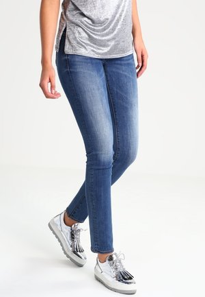 Jeans ajustados azules con un acabado desgastado, combinados con una blusa plateada brillante y zapatillas metálicas con detalles de flecos.