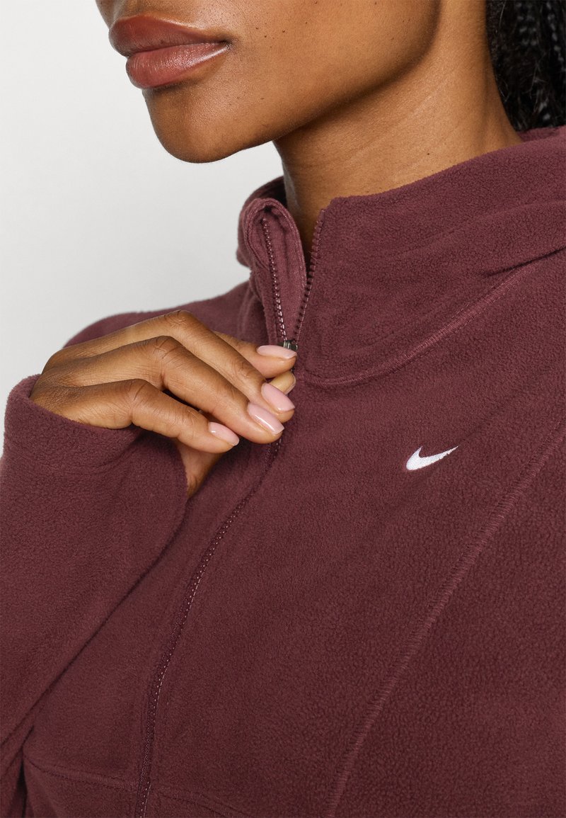 Giacca in pile bordeaux con zip, colletto alto e logo Nike bianco. Il tessuto sembra morbido e strutturato, evidenziando una vestibilità aderente.