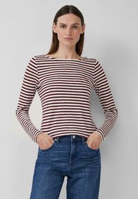 Vrouw met steil bruin haar, gekleed in een lange mouwen kastanjebruin en witte gestreepte top en blauwe jeans, met haar handen in haar zakken.