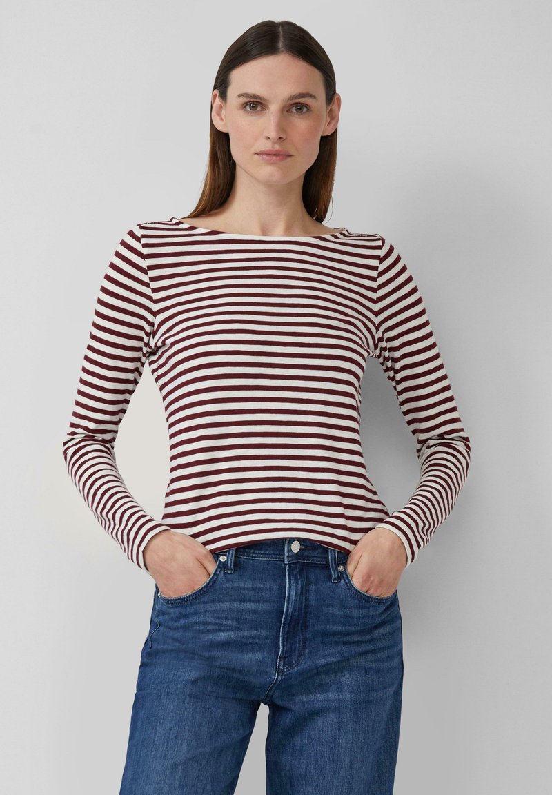 Vrouw met steil bruin haar, gekleed in een lange mouwen kastanjebruin en witte gestreepte top en blauwe jeans, met haar handen in haar zakken.