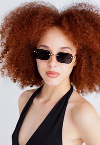 Óculos de sol retangulares pretos com uma moldura prata fina, usados por uma pessoa com cabelo encaracolado vermelho e sardas, contra um fundo neutro.