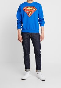 Felpa blu con un grande logo di Superman rosso e giallo, abbinata a jeans in denim scuro e sneakers bianche. Design semplice e casual.