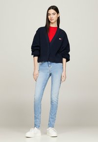 Cárdigan de punto azul marino con cuello en V y cierre de botones, combinado con una camisa roja, jeans de un tono azul claro y zapatillas blancas.