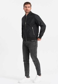 Veste bomber noire avec fermeture éclair sur le devant, poignets côtelés et poche sur la poitrine gauche. Associée à un jean foncé déchiré et des baskets blanches.