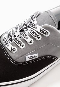 Gros plan d'une basket Vans noire et grise avec semelle blanche, œillets blancs et lacets blancs imprimés à plusieurs reprises avec le logo "Vans" en noir.