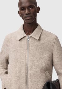 Beige uldblandingsjakke med lynlås foran, krave og sidelommer. Teksturen er blød og let struktureret, med et rent design.