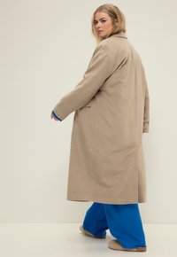 Abrigo largo beige con un ajuste relajado, con cuello y bolsillos laterales, combinado con pantalones sueltos azules y zapatillas de casa de felpa beige.