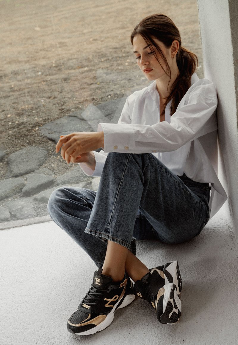 Svarta och gyllene träningsskor med en kraftig sula, texturerad mesh-överkonstruktion och snörningsdesign, tillsammans med ljusblå denimjeans och en vit skjorta.