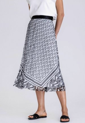 Femme portant une jupe midi plissée avec un motif géométrique noir et blanc, un haut blanc et des sandales plates noires, debout devant un fond uni.