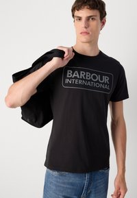Tricou din bumbac negru, cu un logo proeminent gri "BARBOUR INTERNATIONAL". Modelul poartă o geantă de umăr neagră.