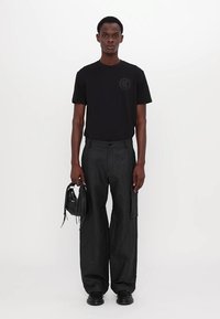 Camiseta de algodón negra con un logotipo redondo, combinada con pantalones negros holgados que tienen un bolsillo lateral. El modelo sostiene un pequeño bolso negro.