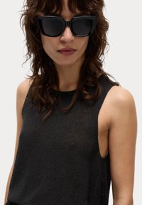 Top negro sin mangas de punto con un acabado texturizado, combinado con unas gafas de sol negras de gran tamaño con una forma angular audaz.