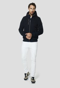 Marineblauer Kapuzenpullover mit Reißverschluss, Fronttasche, verstellbarer Kapuze und kontrastierenden Reißverschlüssen, kombiniert mit weißen Hosen und dunklen Sneakern. Glatte Stoffstruktur.