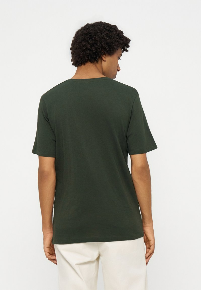 Persona con cabello rizado y corto de espaldas, vistiendo una camisa de manga corta verde oscuro y pantalones de color claro sobre un fondo liso.