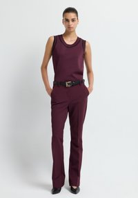 Femme se tenant debout les mains dans les poches, portant un top sans manches maroon et un pantalon assorti, avec une ceinture noire et des chaussures noires à talons pointus.