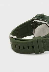 Πράσινο λουράκι ρολογιού από καουτσούκ με υφή, ασημένια αγκράφα και πίσω μέρος κάσας από ανοξείδωτο ατσάλι που δείχνει τις λεπτομέρειες αντοχής στο νερό και το λογότυπο.