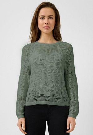 Femme portant un pull en maille vert à manches longues avec un motif géométrique en dentelle et un pantalon noir, debout devant un fond clair uni.