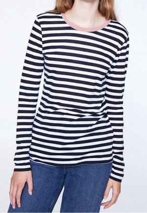 Femme portant un t-shirt à manches longues à rayures horizontales noires et blanches avec un col rose et un jean bleu, debout devant un fond uni.
