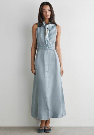 Femme portant une robe sans manches bleu clair boutonnée avec une ceinture à la taille, un foulard à motif autour du cou et des chaussures plates bleues, debout contre un mur uni.