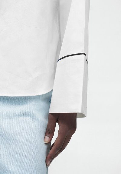 Main à la peau foncée reposant à côté d'un pantalon en denim bleu clair et d'une chemise blanche à manches longues avec un large poignet orné d'un détail de bordure noire.