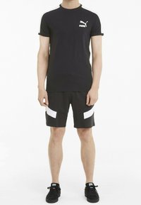 Man wearing black Puma t-shirt, black shorts with white accents, and black Puma sneakers, standing against a white background.