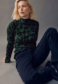 Zöld bordázott garbó fekete leopárdmintával, sötét farmernadrággal párosítva. A modell rövid szőke hajjal, minimális kiegészítőkkel ül.