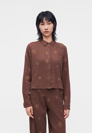 Woman wearing a brown button-up long-sleeve shirt and matching pants with yellow sun and star patterns on a plain white background.