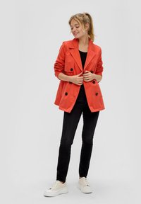 Oranje dubbelgebreide blazer met zwarte knopen, relaxte pasvorm en opgerolde mouwen, gecombineerd met zwarte jeans en witte platform sneakers.