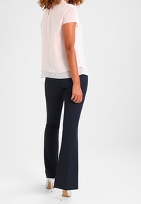 Mujer con cabello rizado que lleva una blusa de manga corta color rosa pálido, pantalones oscuros de campana y zapatos de tacón blanco, de pie de perfil.
