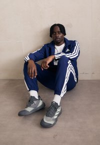 Σακάκι σπορ σε ναυτικό χρώμα με λευκές ρίγες, φερμουάρ και ψηλό γιακά, ελαστικοί καρποί; γκρι αθλητικά παπούτσια με υφασμάτινη σχεδίαση και λευκή σόλα.