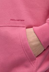 Main dans la poche avant d'un sweat-shirt rose Hollister avec un logo de marque brodé visible au-dessus de la poche.