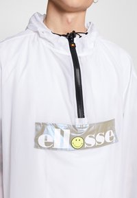 Vit vindjacka med halvdragkedja, som har en glänsande silverfärgad logotyp med svart text och ett gult smiley-ansikte i mitten.