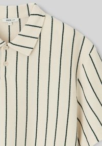 Polo beige avec des rayures verticales vert foncé, présentant un col et trois boutons. Tissu doux et texturé avec une coupe ample.
