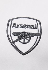 Logo del club di calcio Arsenal ricamato in nero su tessuto bianco, che presenta un cannone con una ruota all'interno del contorno di uno scudo.