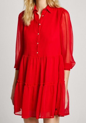 Femme portant une robe rouge vif boutonnée, à étages, avec des manches longues semi-transparentes et un col, montrée du cou jusqu'à la mi-cuisse.