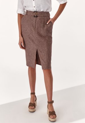 Femme portant une jupe crayon marron ceinturée avec une fente frontale, une chemise blanche aux manches retroussées et des sandales compensées marron avec des brides à la cheville.