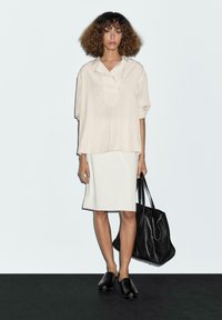 Lichtbeige oversized shirt met een kraag, witte aansluitende rok, zwarte leren handtas en zwarte instap-schoenen met studs.