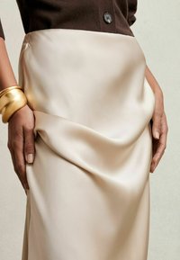 Falda de satén beige con un diseño drapeado, combinada con un suéter de punto marrón. Una pulsera de oro adorna una mano, destacando la textura y el contraste de colores.