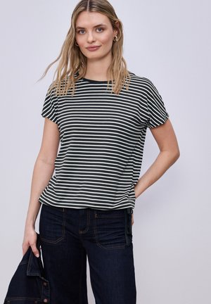 Frau mit langem blondem Haar, die ein kurzärmeliges schwarz-weiß gestreiftes Shirt und dunkle Jeans trägt, hält eine dunkle Jeansjacke in der linken Hand.