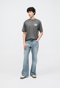 Grå bomullst-shirt med tryckt logotyp, kombinerad med ljusblå utsvängda jeans. Modellen bär vita sneakers med bruna detaljer.